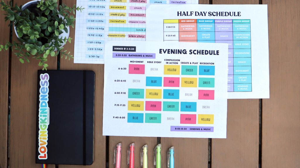 A potted plant sits on a table next to a black notebook with a "lovingkindess" sticker. Three papers neatly overlap each other, each with a different sample VBS schedule. Below the schedules are five highlighters of different colors.