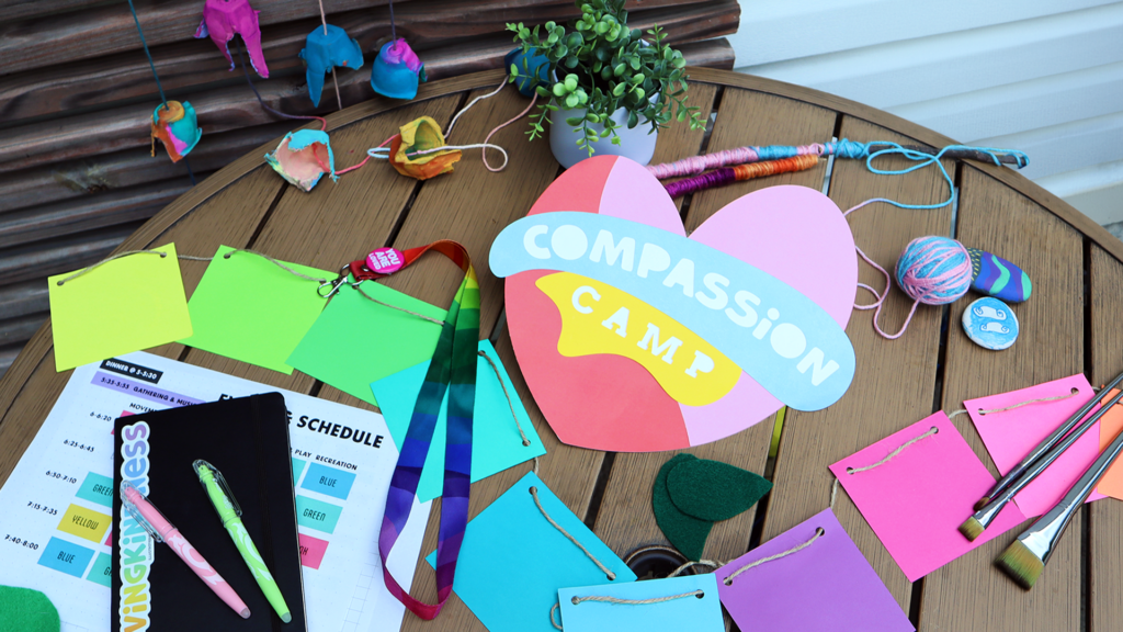 Outdoor wooden table against a white and brown wall. The table is covered with vibrant craft supplies and organization tools. In the middle is a large paper heart, it's the logo for Compassion Camp.