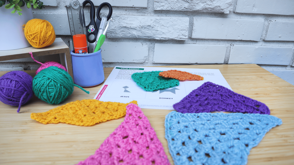 A desk. On the desk are tangram shapes crocheted in different colors and a Storytelling with Shapes puzzle page. On the back-left side of the desk is a plant in a white pot on top of a light wooden stand. Next to and below the plant are colorful balls of yarn and a crochet hook The background is a white brick wall.