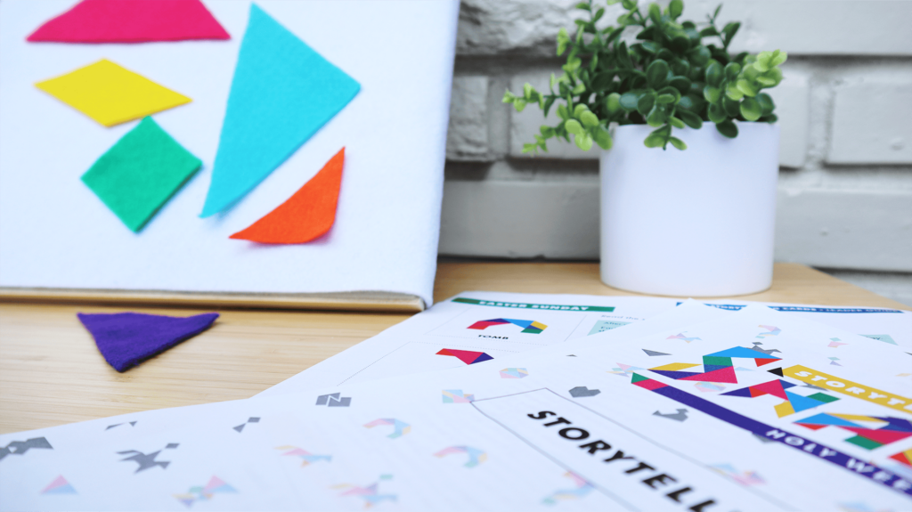 A close up of Storytelling with Shapes  adapted for use on a felt board. Colorful tangram shapes hang on the board and one lays on the table. In the background there is a plant in a white pot and a white brick wall.